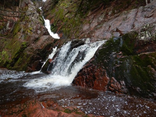 A cascading waterfall flows over moss-covered rocks in a lush, green forest setting.