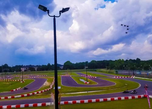 A go-kart track with racers, under a cloudy sky, as jets fly overhead in formation.