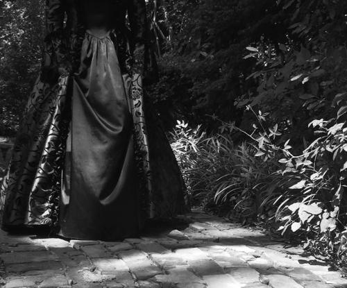 A figure in a dark, ornate dress stands on a cobblestone path surrounded by lush greenery, captured in black and white.