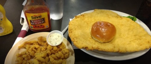 A large breaded fried dish with a bun, accompanied by a side of fried shrimp and ketchup on a table.