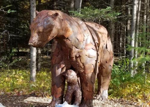 A large bear sculpture stands beside a smaller bear, both made of textured metal, set in a forested area.