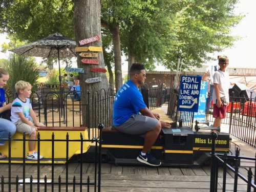 A child rides a pedal train while another child and an adult wait nearby in a sunny outdoor setting.