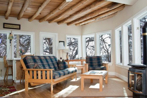 Cozy living room with wooden beams, large windows, and comfortable seating, featuring a small table and a wood stove.