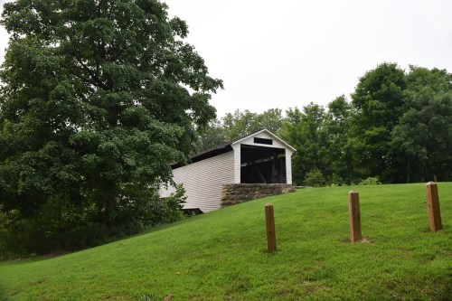 A white covered bridge sits on a grassy hill, surrounded by trees under a cloudy sky.