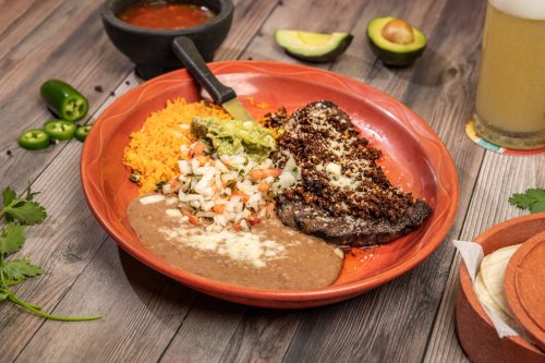 A vibrant plate of Mexican food featuring grilled steak, rice, guacamole, pico de gallo, and refried beans.