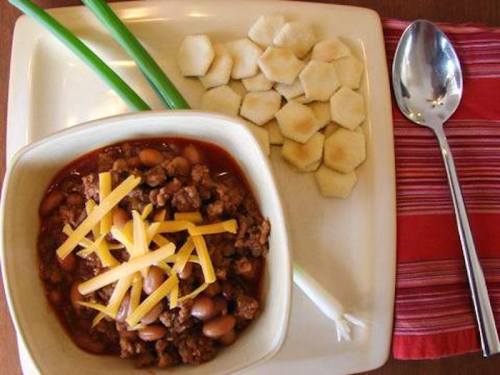 A bowl of chili topped with cheese, served with sliced crackers and green onions on a plate.