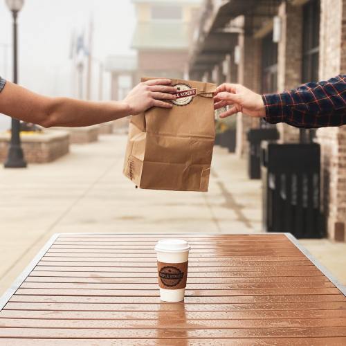 A hand passes a brown paper bag over a table with a coffee cup in the foreground, set in a foggy outdoor area.