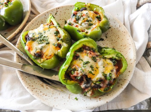 Stuffed green bell peppers topped with melted cheese and herbs, served on a speckled plate with utensils.