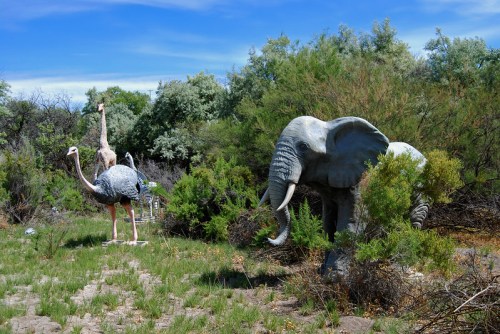 A lifelike elephant statue and two ostrich sculptures among green shrubs and trees under a blue sky.