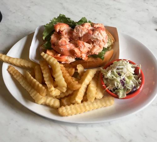 A plate featuring a lobster roll on a bun with lettuce, crinkle-cut fries, and a small bowl of coleslaw.
