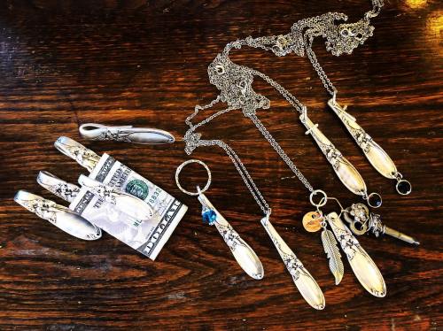A collection of silver jewelry pieces, including necklaces and keychains, arranged on a wooden surface with a dollar bill.