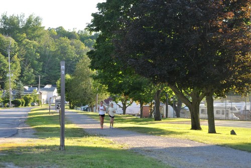Two people jog along a tree-lined path beside a quiet street in a rural area.