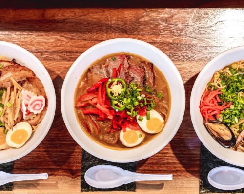 Three bowls of ramen with various toppings, including sliced meat, vegetables, and soft-boiled eggs, on a wooden table.