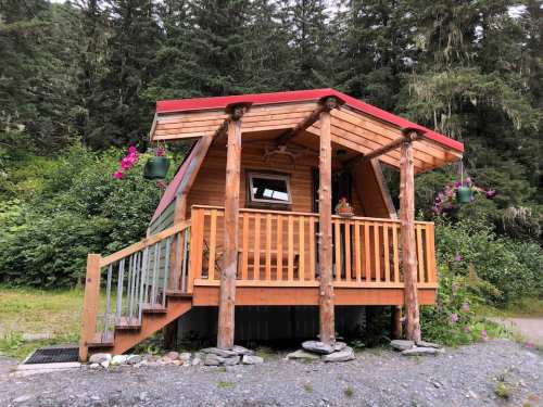 A cozy wooden cabin with a red roof, surrounded by greenery and flowers, featuring a porch and steps.