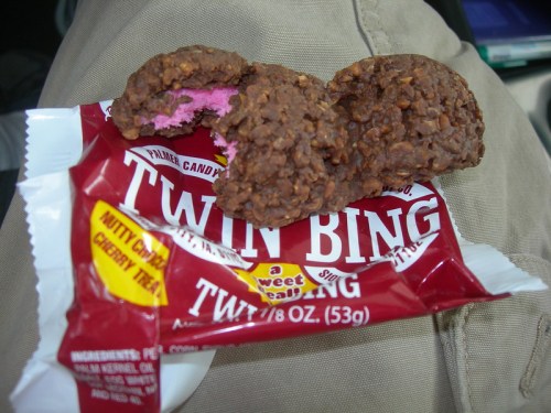 A close-up of a Twin Bing candy bar, partially unwrapped, with a few pieces showing the chocolate and pink filling.