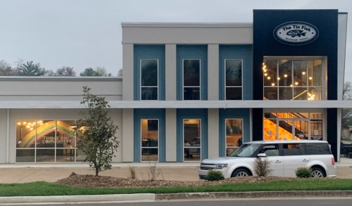 Exterior view of a modern building featuring "The Tin Fish" sign, with large windows and a parked white car in front.