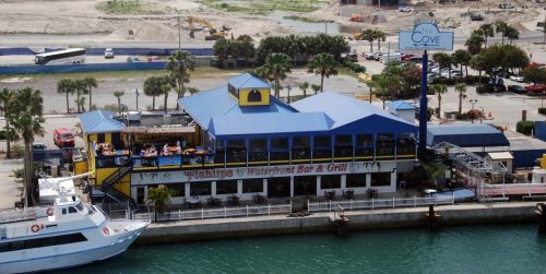 A waterfront bar and grill with a blue roof, surrounded by palm trees and construction in the background.