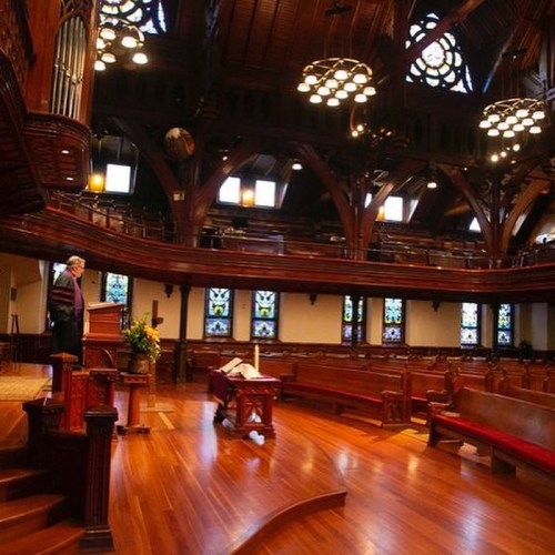 A spacious, ornate church interior with wooden beams, stained glass windows, and a speaker at the podium.