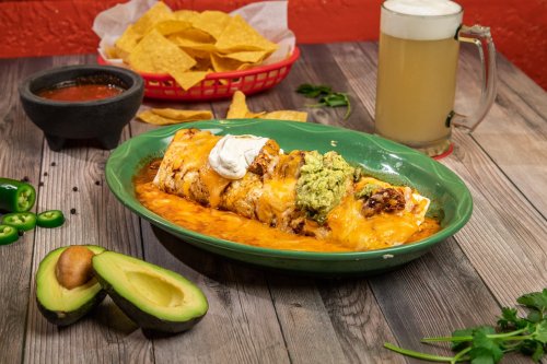 A plate of enchiladas topped with cheese, guacamole, and sour cream, served with chips, salsa, and a beer.