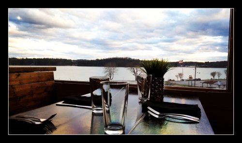 A table set for dining with glasses and utensils, overlooking a lake and cloudy sky in the background.