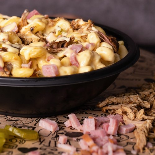 A bowl of creamy macaroni and cheese topped with shredded meat and diced ham, served on a patterned surface.