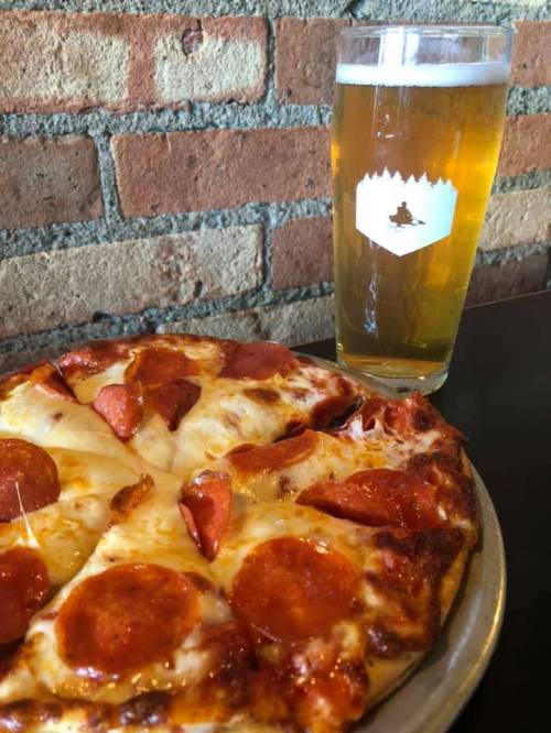 A slice of pepperoni pizza on a plate next to a tall glass of golden beer against a brick wall.