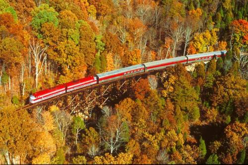 A red train crosses a wooden trestle bridge surrounded by vibrant autumn foliage.
