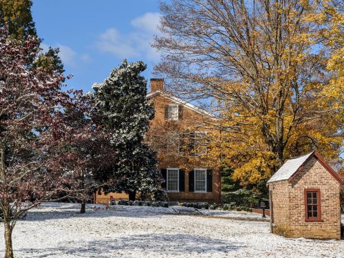 A historic brick house surrounded by trees with autumn leaves and a light dusting of snow on the ground.