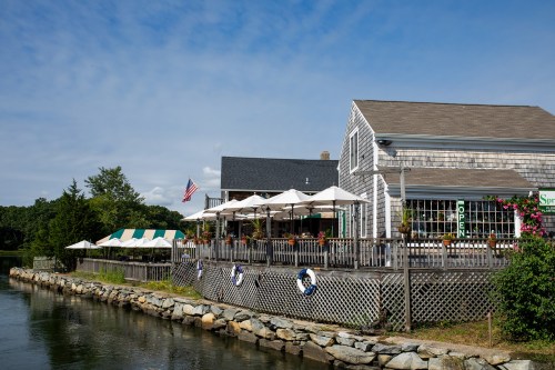 A waterfront restaurant with outdoor seating, umbrellas, and an American flag, surrounded by greenery and a calm waterway.