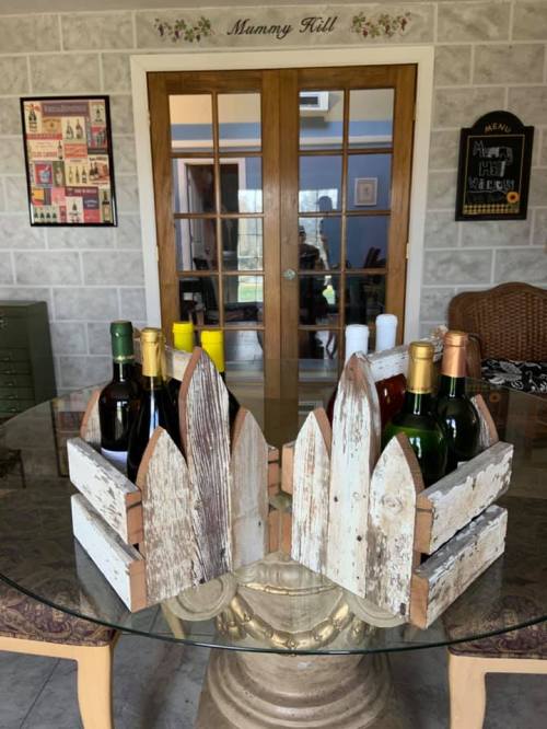 A rustic wine holder made of weathered wood, displaying several bottles of wine on a glass table in a cozy room.