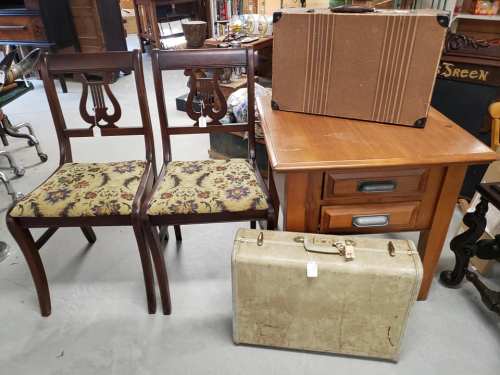 Two wooden chairs with floral seats, a vintage suitcase, and a wooden side table with drawers in an antique shop.
