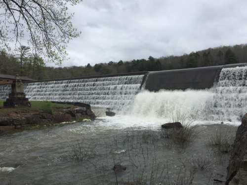 A dam with cascading water flows over a stone structure, surrounded by greenery and a cloudy sky.