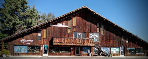 A rustic wooden building with a sloped roof, featuring large windows and a balcony, surrounded by trees.