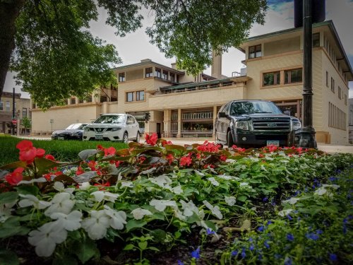 A view of a building with a modern design, surrounded by colorful flower beds and parked cars.
