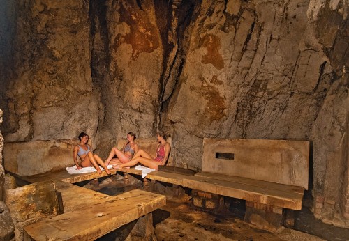Three women relax in a rustic stone sauna, sitting on benches in a cave-like setting.