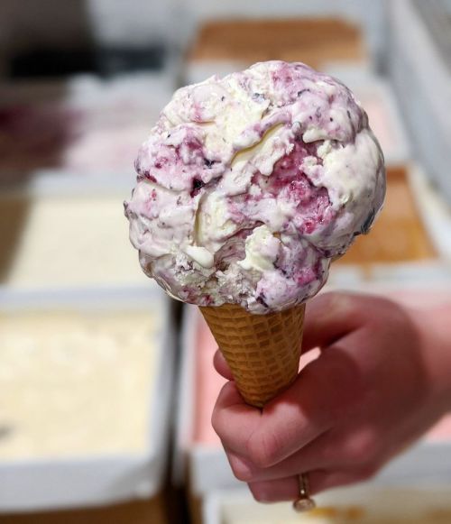 A hand holding a large ice cream cone with swirls of purple and white flavors against a blurred background of ice cream tubs.