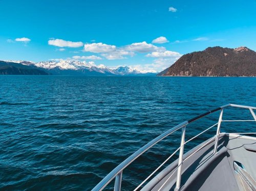A boat's bow cuts through calm blue waters, with snow-capped mountains and a forested shoreline in the background.