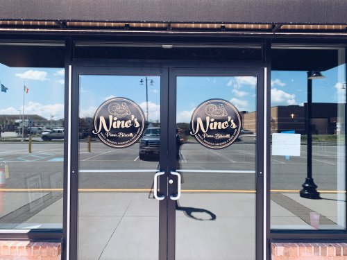 Double glass doors with round signs reading "Nino's Pizza & Brew" in a storefront. Clear sky visible in the background.