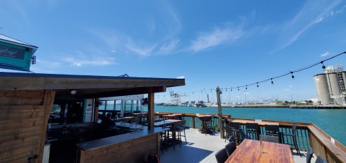 A sunny waterfront view from a deck with seating, overlooking a harbor and industrial buildings in the distance.