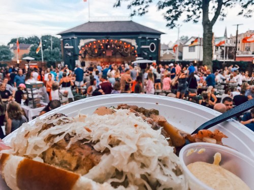 A close-up of a plate with a sausage and sauerkraut, with a lively outdoor crowd and stage in the background.