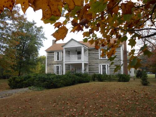 A historic wooden house with a porch, surrounded by autumn leaves and trees in a serene landscape.