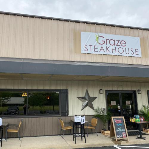 Exterior of Graze Steakhouse with a sign, outdoor seating, and a menu board displaying takeout options.