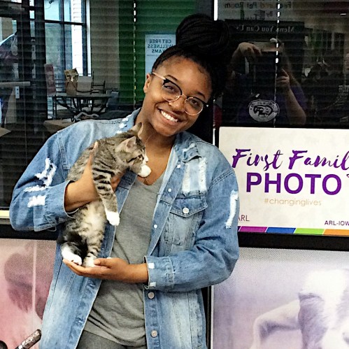A smiling person holds a gray tabby kitten in a bright, indoor setting with colorful signs in the background.