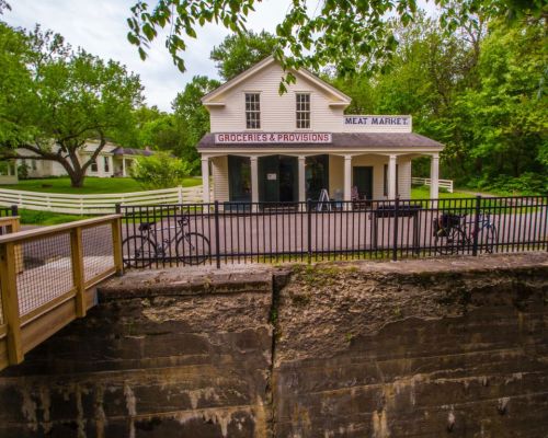 A historic building labeled "Groceries & Provisions" and "Meat Market," surrounded by greenery and a wooden fence.