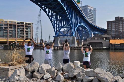 Four people stand by a river, forming the letter "O" with their arms, under a blue bridge.