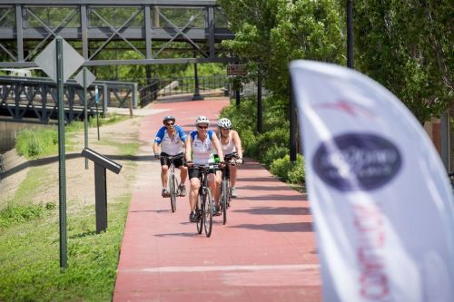 Three cyclists ride along a paved path beside a river, with greenery and a flag in the foreground.