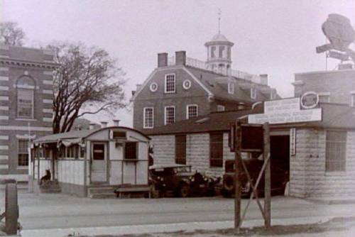 Historic black-and-white photo of a building with a trolley stop and vintage cars parked outside.