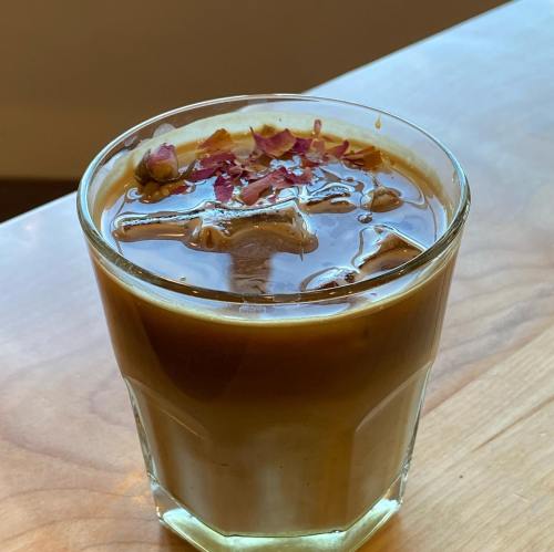 A glass of iced coffee topped with rose petals, sitting on a wooden surface.