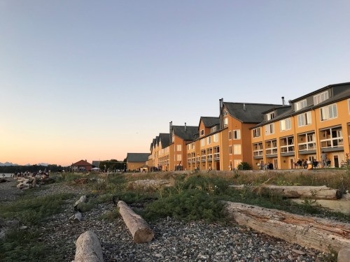 A beachfront hotel with yellow walls at sunset, surrounded by logs and greenery along the shore.