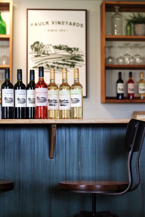 A wooden bar with various wine bottles displayed, framed vineyard artwork in the background, and a chair in the foreground.
